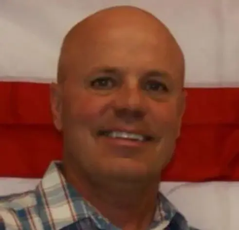 A bald man with a checkered shirt and a flag behind him.